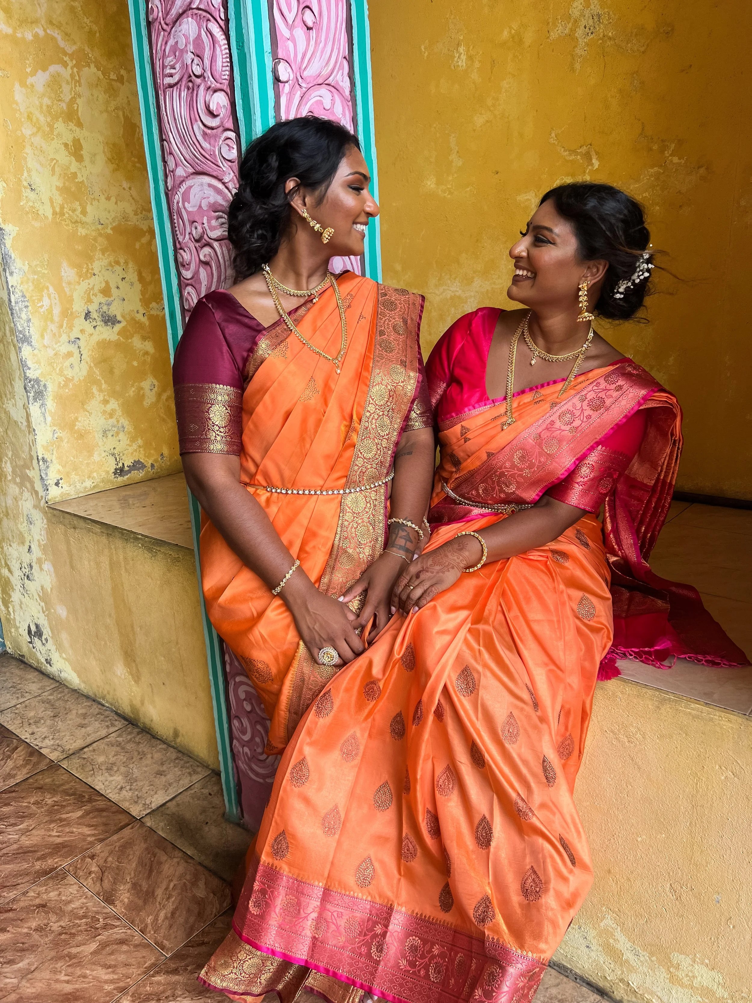 Two women in traditional orange sarees sitting against a yellow wall.